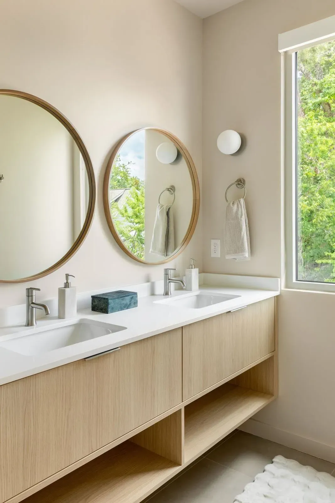 Primary bath with a double wood vanity, round mirrors, and a window to the trees